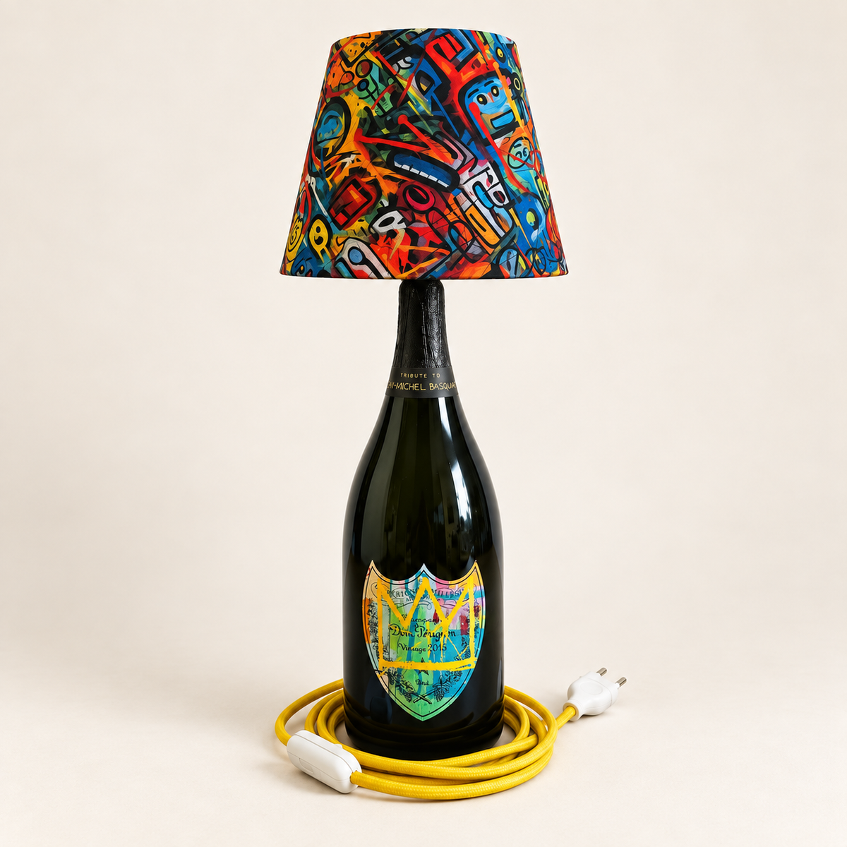 Dom Pérignon bottle lamp with yellow Basquiat crown, colorful fabric shade, and yellow fabric-covered cord, switched off.